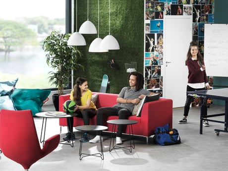 A modern, collaborative workspace featuring a casual seating area with red sofas and black tables. Two people are sitting on the sofas engaged in conversation, while another person stands in the background near a flip chart, actively presenting. The room is designed with a vibrant and creative atmosphere, including a green plant wall, pendant lights, and a large window allowing natural light to enter. The setting suggests an informal yet productive environment, ideal for teamwork and brainstorming sessions.