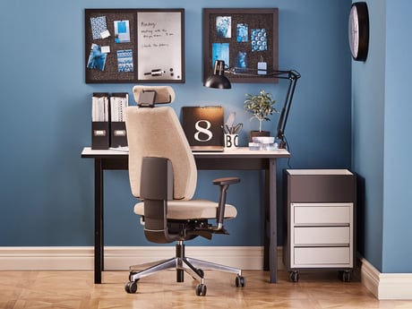 A stylish home office setup featuring a beige ergonomic chair with a headrest, positioned in front of a black desk. The desk is adorned with neatly organised files, a desk lamp, and decorative elements, including a plant and a calendar. The backdrop includes a vibrant blue wall and a pinboard displaying notes and photos, creating a modern and productive workspace.