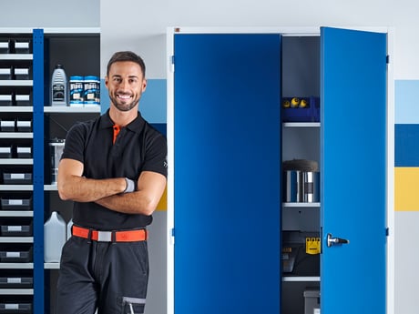 A man leaning against a tall metal cabinet. The cabinet has a light grey frame and blue doors. One door is ajar showing tools stored inside. In the background there is metal shelving with small parts boxes and cleaning liquids on the shelves.