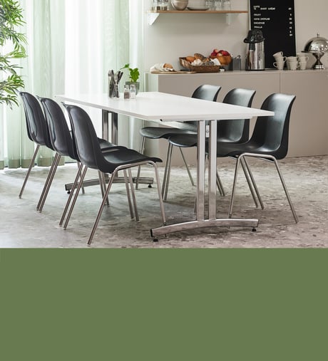 In focus, a rectangular canteen table with a white table top and silver legs surrounded by six black, plastic chairs with silver legs. In the background, an office break area with breakfast buffet.