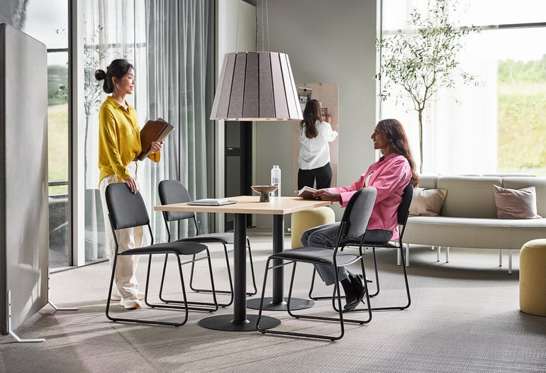 Two women are in a modern office space designed by AJ Products. One woman in a yellow shirt stands near a partition holding folders. Another woman in a pink shirt sits at a table, reading. A third woman is visible in the background, writing on a board.