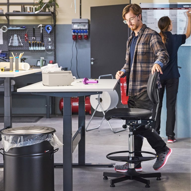 A person stands at a complete height-adjustable workbench beside a draughtsman chair in a warehouse setting.