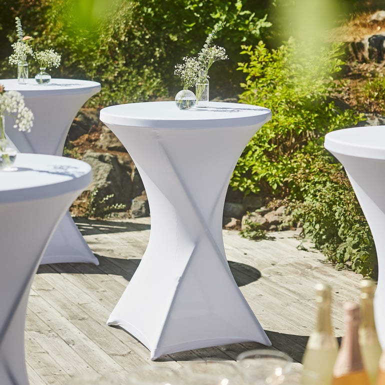 Several round cocktail tables covered in white stretch cover CLARA, arranged outdoors on a wooden deck