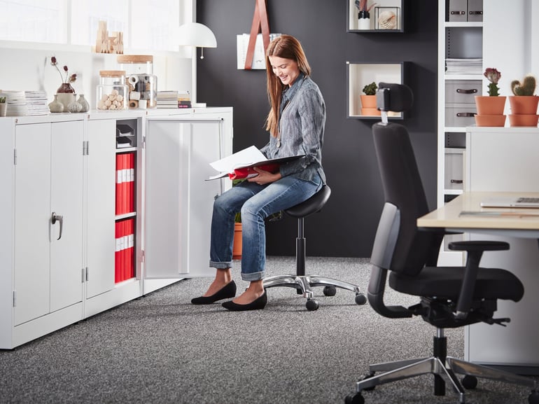 A bright and organised office setup featuring a woman sitting on a black ergonomic saddle chair, reviewing documents. The background includes sleek white storage cabinets filled with vibrant red binders and decorative jars, along with potted plants adding a natural touch. The workspace blends functionality and style with a professional yet inviting atmosphere.