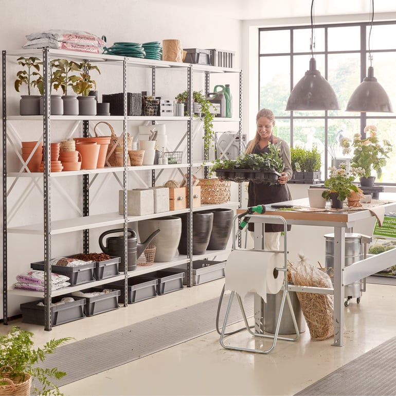 Blomsterhandler bærer en bakke med planter i en butik med metalhylder