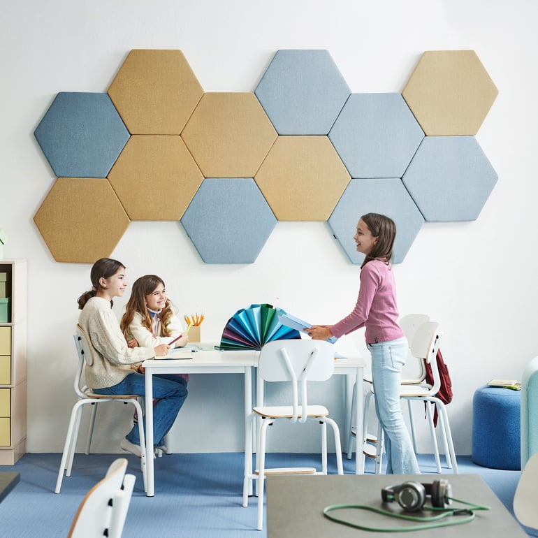 Tre barn i ett klassrumsmiljö sitter vid ett vitt bord med vita stolar. Två barn sitter och ett står upp. På väggen bakom dem sitter hexagonformade ljudabsorbenter i blått och gult. Rummet har en modern och färgglad design med organiserade skrivbordstillbehör.