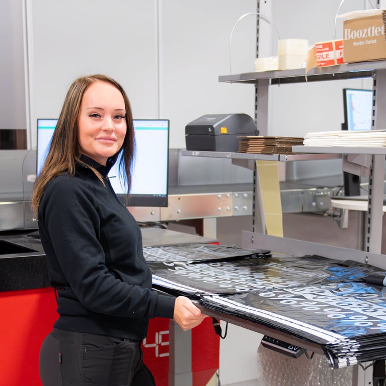 Woman working at a packing station. She is standing at a workbench with a computer screen and packing bags on the worktop.