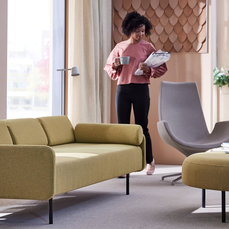 cozy and modern living space in natural light.  In the foreground, there is a contemporary couch with a muted olive-green fabric. In the background a woman walks holding a cup and a pile of papers
