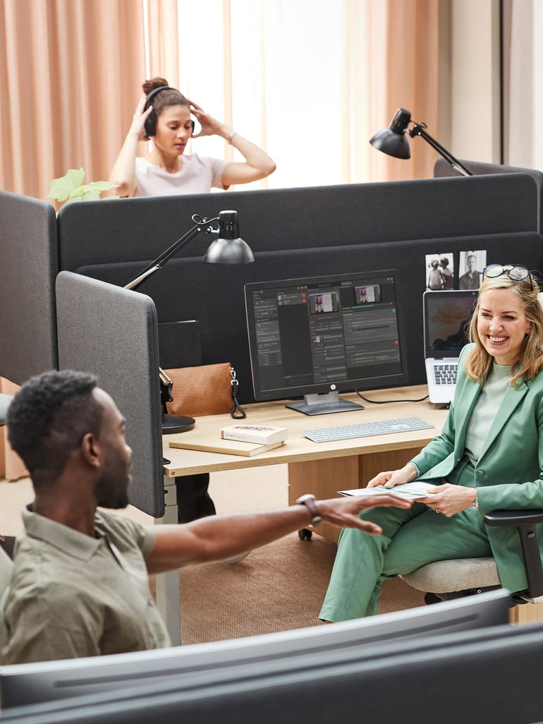 a modern co-working space with three individuals. Two women are seated at individual workstations separated by cubicle dividers, while a man appears to be reaching across the dividers to interact with one of the women. One woman is engaged in a video call or listening to audio. There is an overall atmosphere of collaboration and professional interaction within a shared workplace.