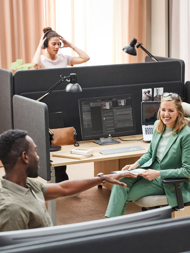 a modern collaborative open office environment where two people interact and in the background another person adjust her headphones 