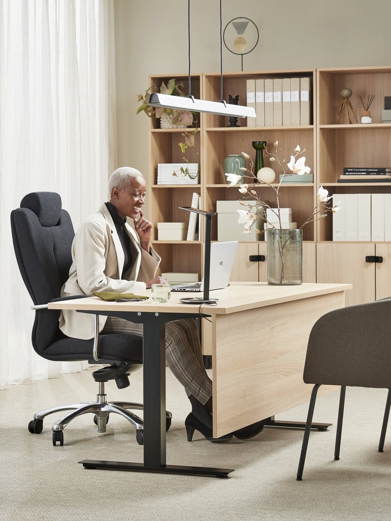 A woman working at a desk in an office sitting in a SHEFFIELD office chair.  She has a laptop open and is looking at it. The desk is made of wood and has a modern design. There is a bookcase with files and decorative items in the background.