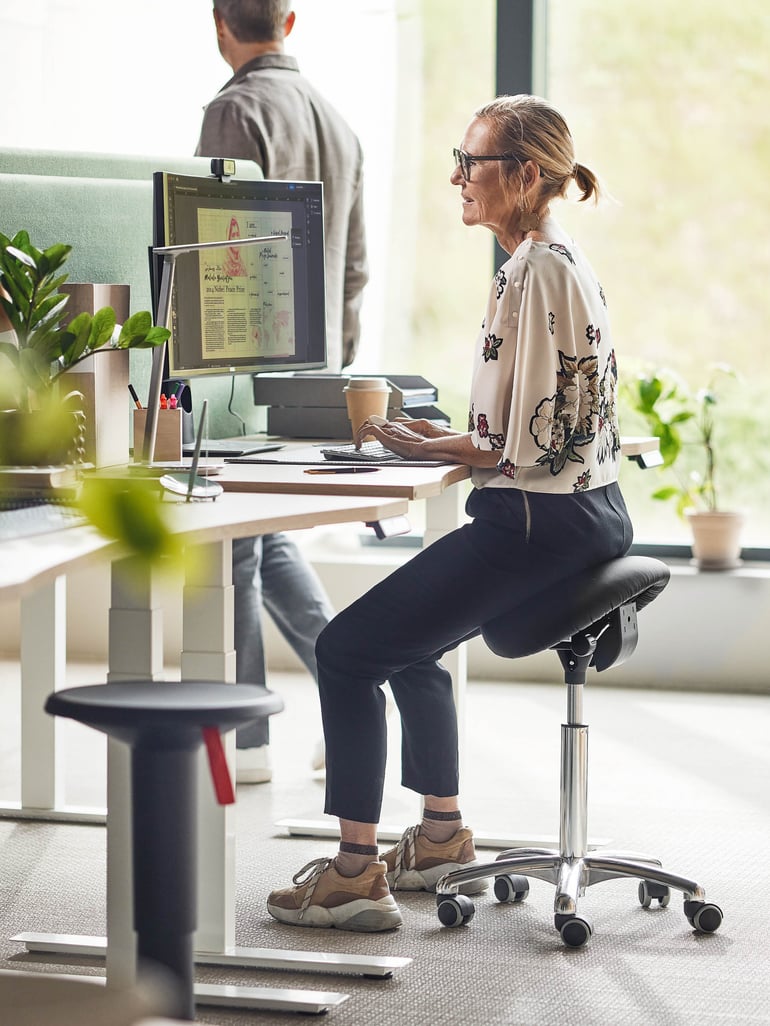 Personen som sitter på en sadelpall med en flexibel stol vid ett skrivbord med en dator.