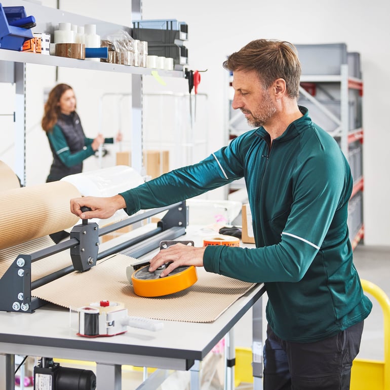 Person som använder en lådkniv vid en förpackningsarbetsstation i en verkstadsmiljö.