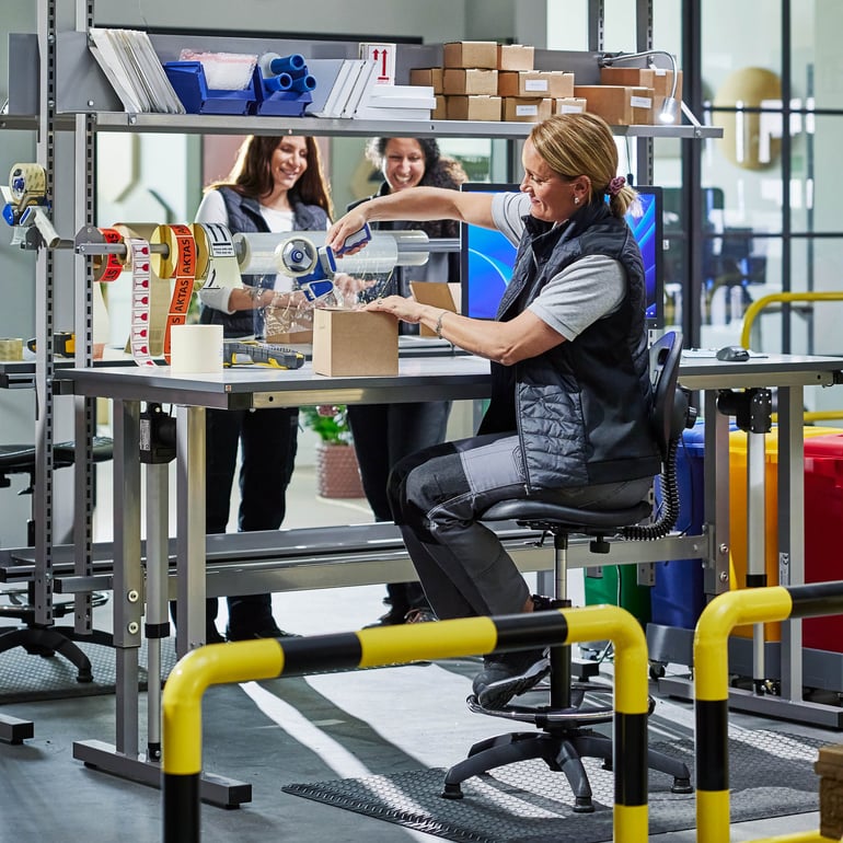 En person använder tejp för att försegla en låda på en höj- och sänkbar arbetsbänk i en verkstad.