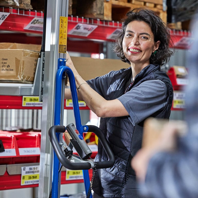 Person som ler direkt mot kameran i en lagermiljö med en handbogserad staplare.