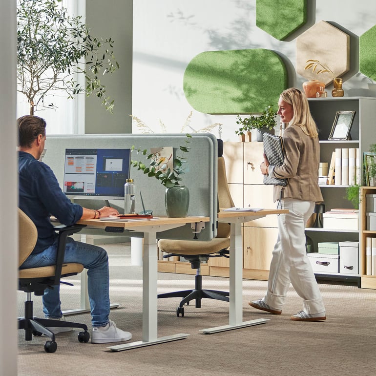 Två personer vid ett skrivbord på ett kontor, med en växt i närheten och en dator på bordet.