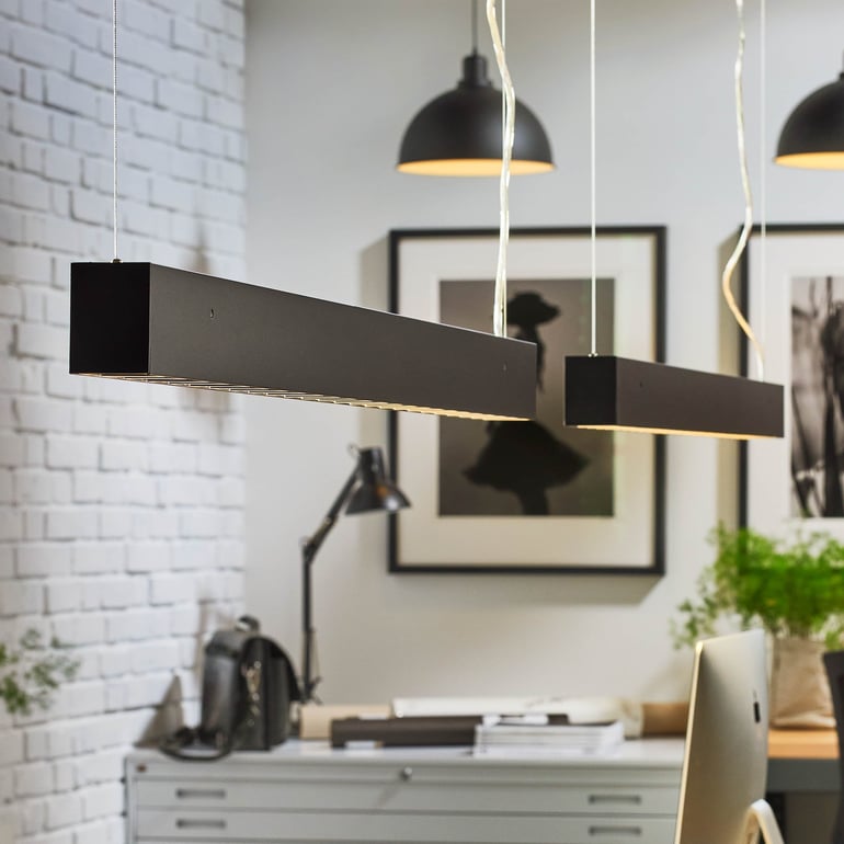 En svart rektangulär taklampa lyser upp ett modernt kontorsutrymme med en växt i närheten.