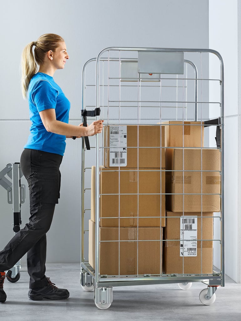 En person knuffar en rullbehållare fylld med lådor i en lagermiljö.