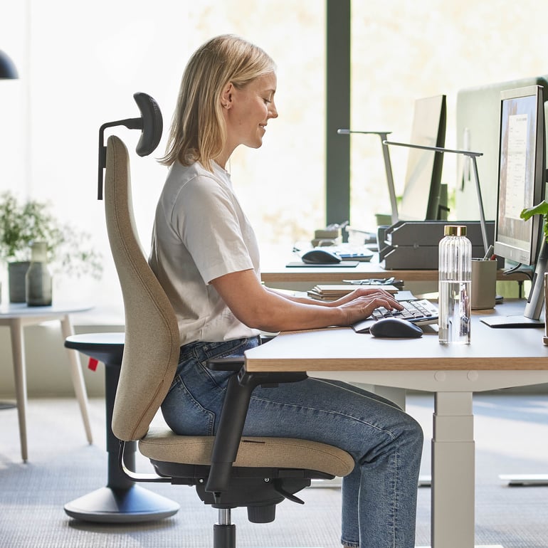 Person som sitter i en kontorsstol vid ett skrivbord med hjälp av en dator, med en växt i närheten.