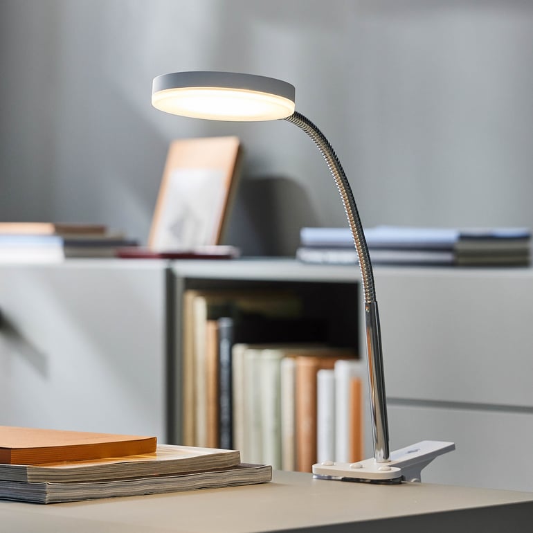 Skrivbordslampa placerad bredvid en trave böcker på ett skrivbord. Mjukt ljus lyser upp området.