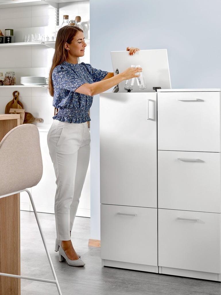 The image shows a woman in a modern, clean office kitchen or workspace setting. She is wearing a blue patterned blouse, white trousers, and white heels. The woman is opening the top of a white storage cabinet and placing a clear spray bottle inside. The space features a bright and minimalist design, with white cabinetry, neatly arranged shelves, and natural light streaming in, creating an organised and professional atmosphere.