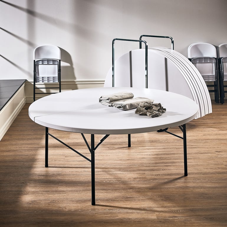 A round foldable white table with black metal legs standing on a smooth wooden floor.  Behind this main table, there's a stack of additional round tabletops, leaning vertically in a table trolley