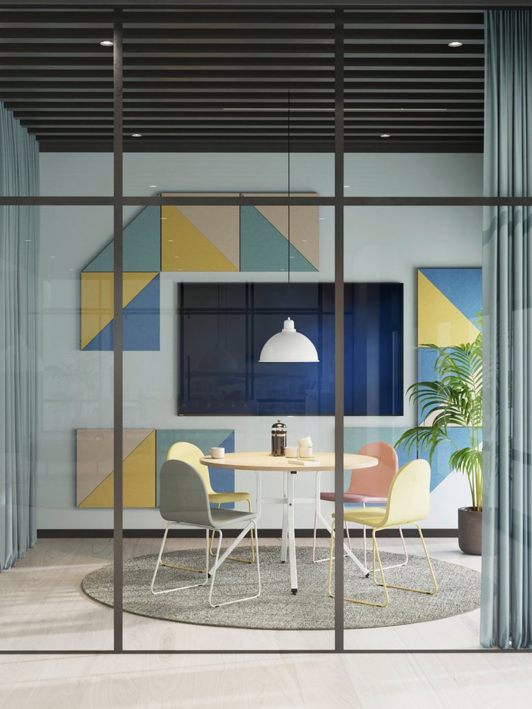 The image shows a contemporary meeting space with a round white table surrounded by four colourful chairs in pastel shades of green, pink, yellow, and grey. The area is enclosed by glass walls, creating an open and airy atmosphere. The wall behind the table features geometric artwork in shades of yellow, blue, and green, complementing the modern decor. A large screen is mounted on the wall, with a white pendant lamp hanging above the table. A round rug defines the seating area, and a lush potted plant adds a touch of greenery. The space exudes a stylish yet functional vibe.