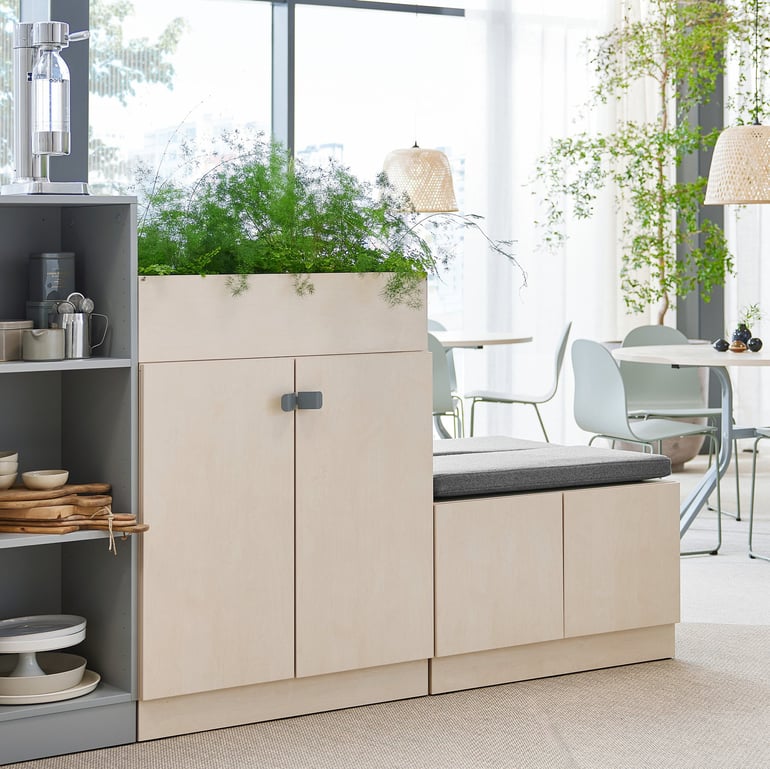 a light-colored QBUS wooden storage unit by AJ Products with two compartments: a two-door cabinet on the left and a cushioned bench with storage on the right. The cabinet has various kitchenware items. On the top of the cabinet, there is a lush green plant adding a touch of nature and freshness,