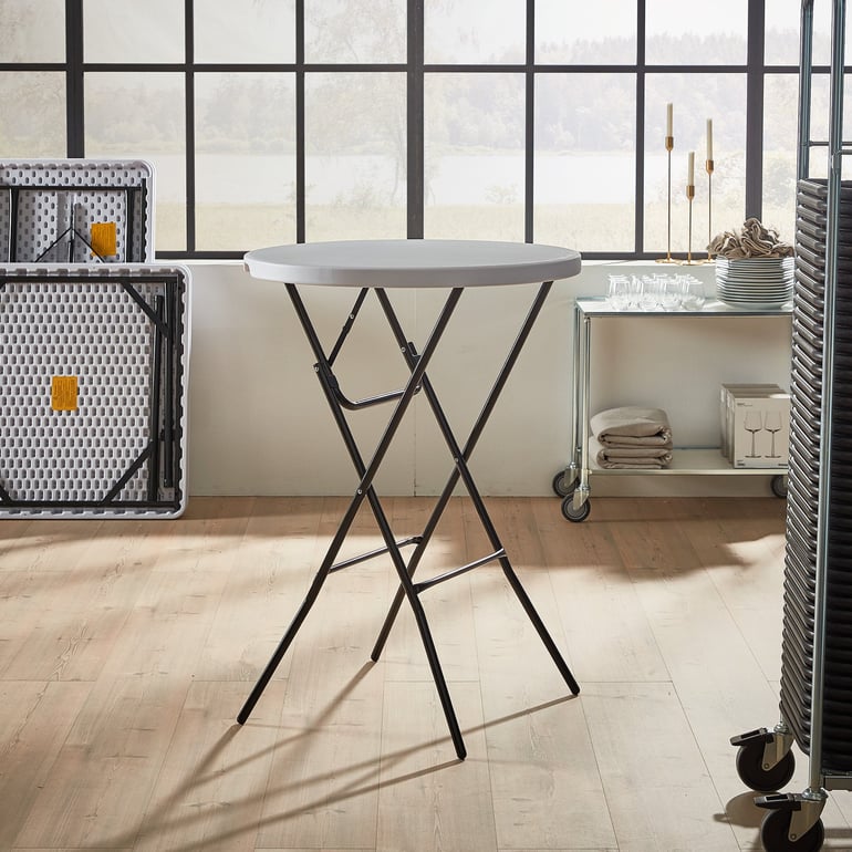 a setting for events and catering with a plastic round folding table with a black metal frame. It's positioned in a bright, airy room with large windows and light-colored wood floors. In the background, there are other folded tables, a serving cart with dishes and linens, and some candlesticks
