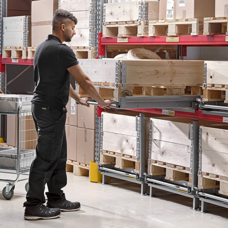 a man working in a large warehouse. He is pulling a wooden pallet using a metal handle. The pallet is part of a system of metal racking and shelving storage.