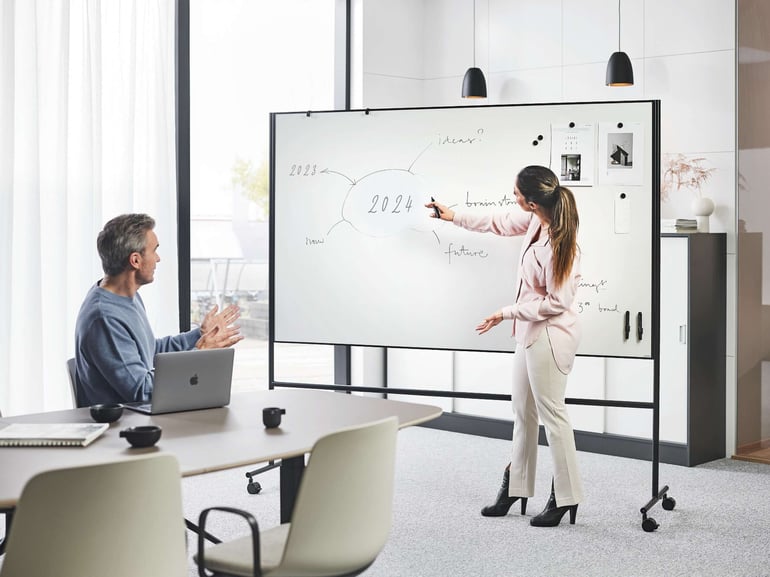 To personer holder et møde. Den ene sidder ned og den anden skriver på et whiteboard med hjul.