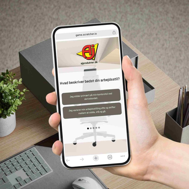 En person holder sin mobiltelefon i hånden. På skærmen ses AJ Produkters stoleguide, som personen er ved at prøve, for at finde den rigtige kontorstol.