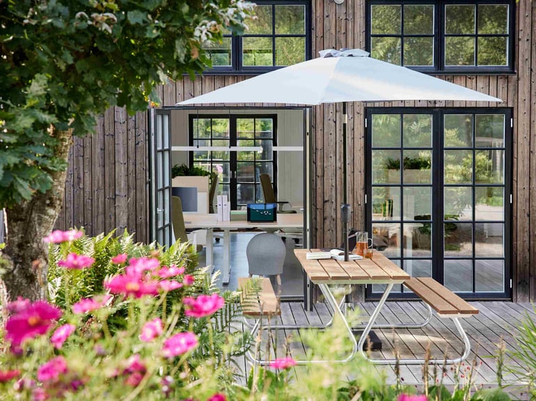 Hyggelige udearealer udenfor nogle kontorlokaler. Der er blomster og træer, borde og bænke og en parasol.