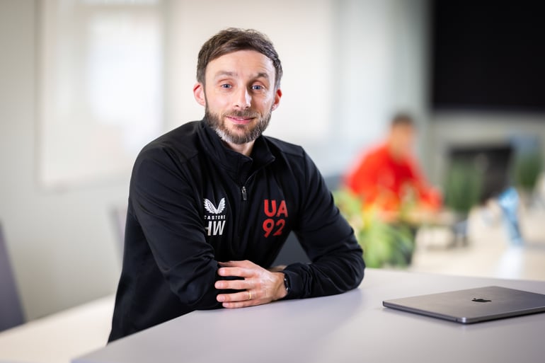 Man in a UA92-branded jacket sitting at a desk, with a laptop in front of him.