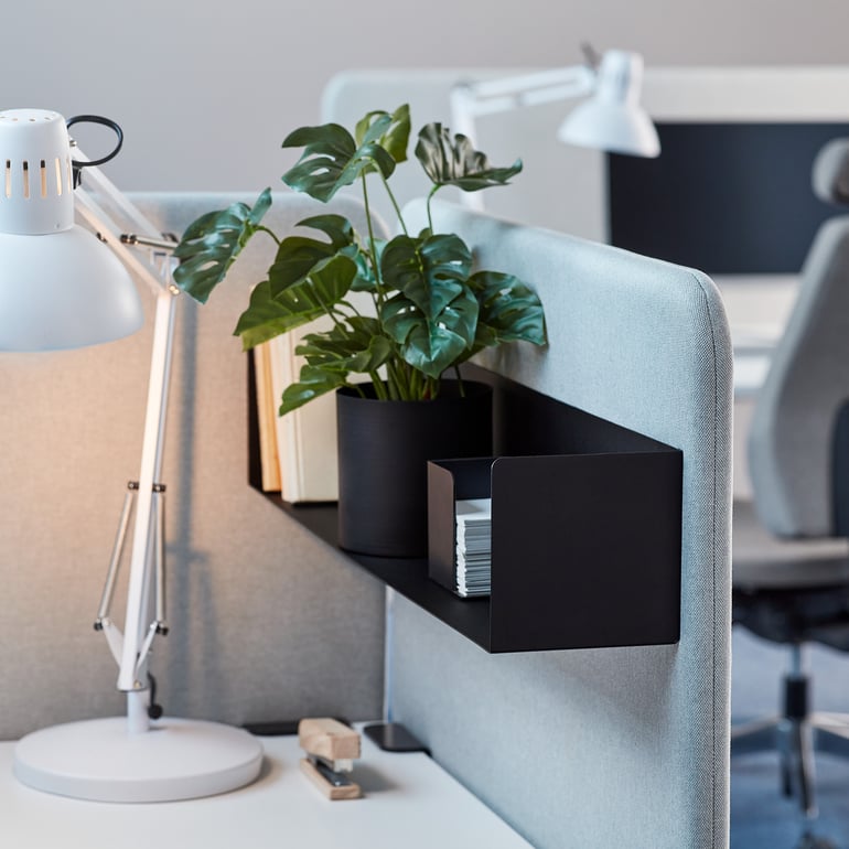 The image showcases a modern office workspace with a partitioned desk. The desk features a white lamp with an adjustable arm, a black metal storage shelf attached to the partition, a potted green plant, and neatly arranged office supplies. In the background, additional workstations with similar setups and office chairs are visible, creating a professional and organised environment. The colour scheme includes neutral tones like grey, white, and black, with the green plant adding a touch of vibrancy.