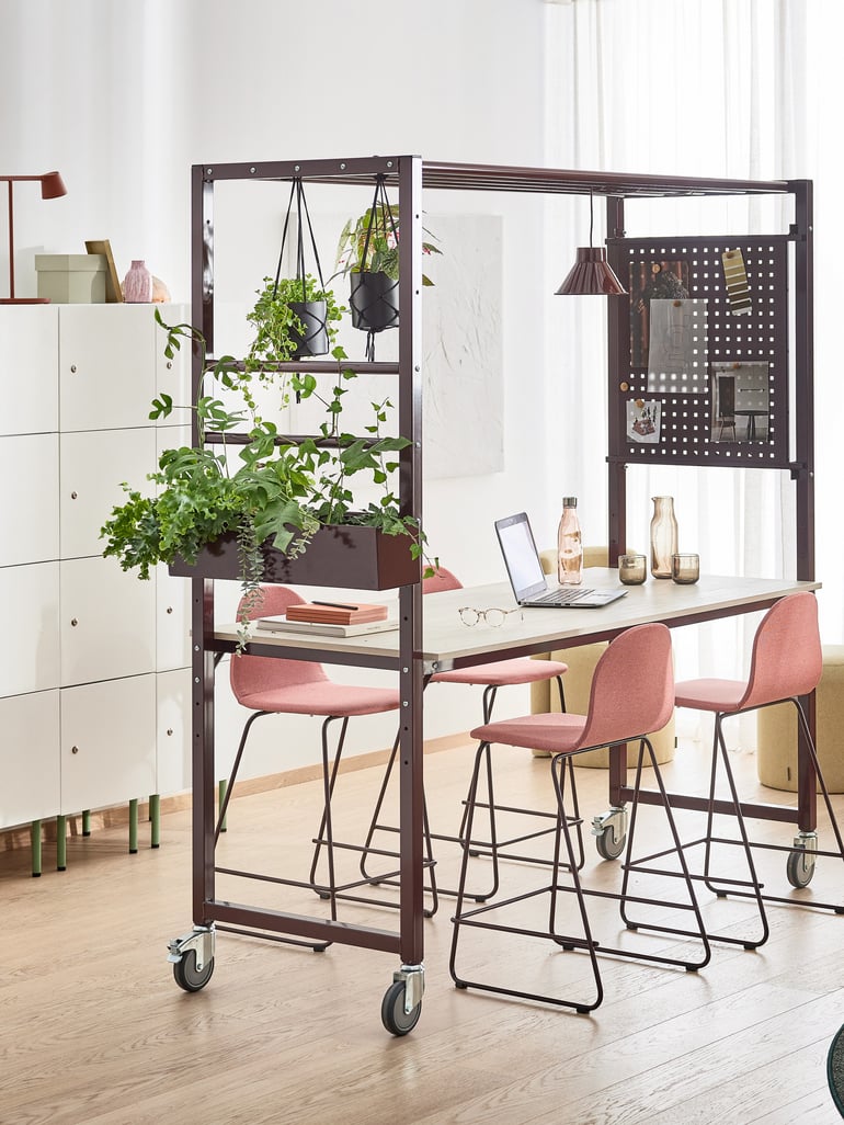 Flexible coworking space with a mobile workstation that consists of a high table with a metal frame on wheels. It is paired with pink upholstered barstools with metal legs. Above the table, there is an integrated structure that holds hanging plants, a pegboard with notes and inspiration materials, and a small pendant lamp for task lighting. The background features white storage cabinets, decorative items, and soft natural light filtering through sheer curtains. 