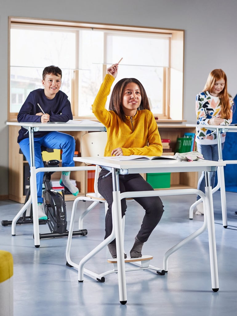 A classroom with a few students where a girl in a yellow sweater sitting at a desk raise her hand to answer a question