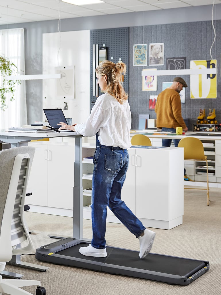 En person går på et gåbånd mens hun arbejder ved at hæve sænkebord på et kontor