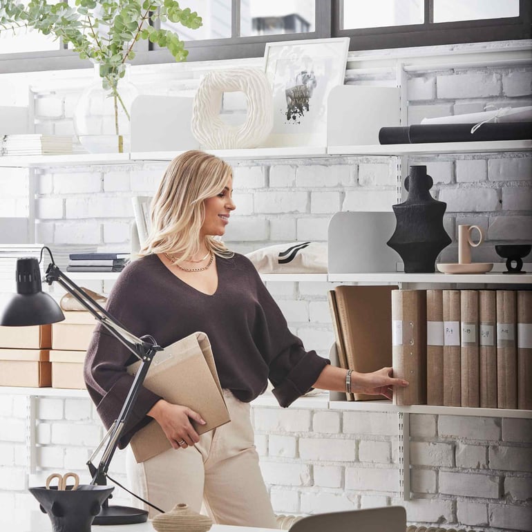 En kvinde på et lyst kontor står med en medarbejderhåndbog under armen ved en vægreol