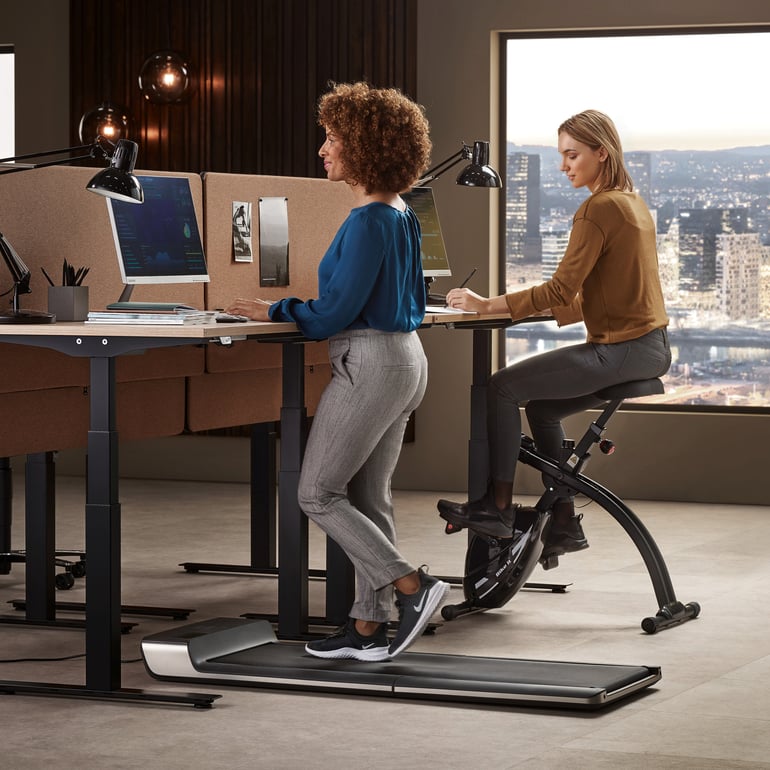 A woman working at a desk on a treadmill and a woman working at a desk on an exercise bike