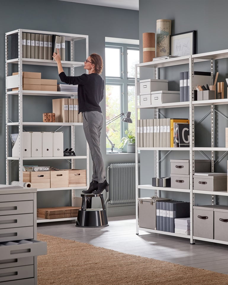 A woman stands on a stool and retrieves a binder from a high office shelf