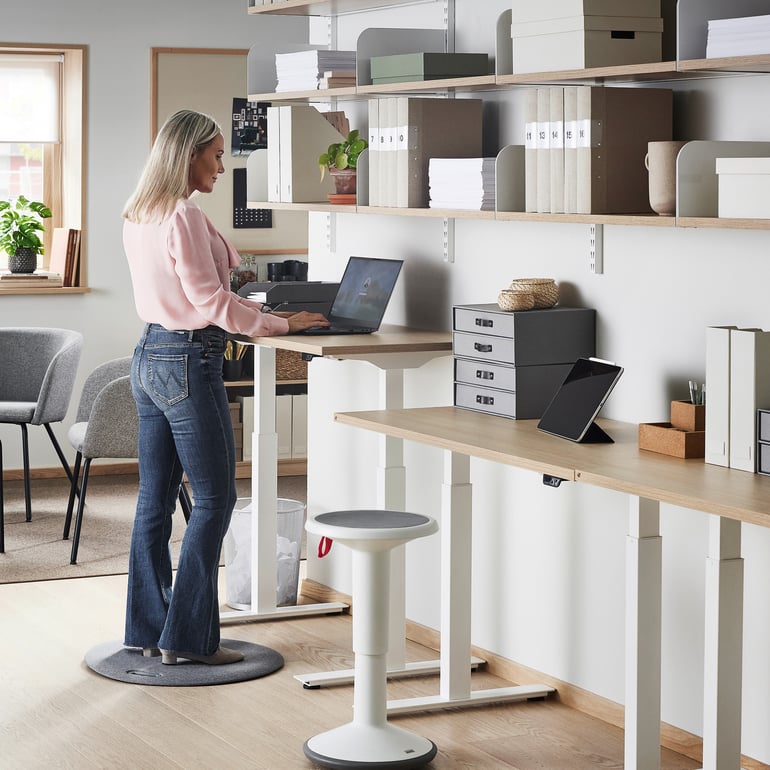 An office with height-adjustable desks with a woman stood on a standing mat at a desk