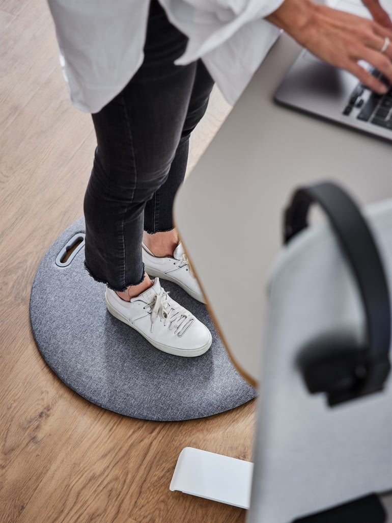 A person is standing on a round workplace mat at a height-adjustable desk and typing on a laptop