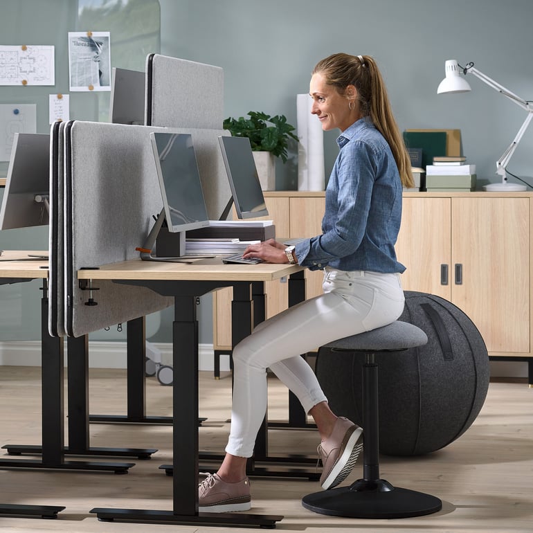 Moteris sėdi ant balansinės aktyvaus sėdėjimo kėdės, prie biuro stalo.