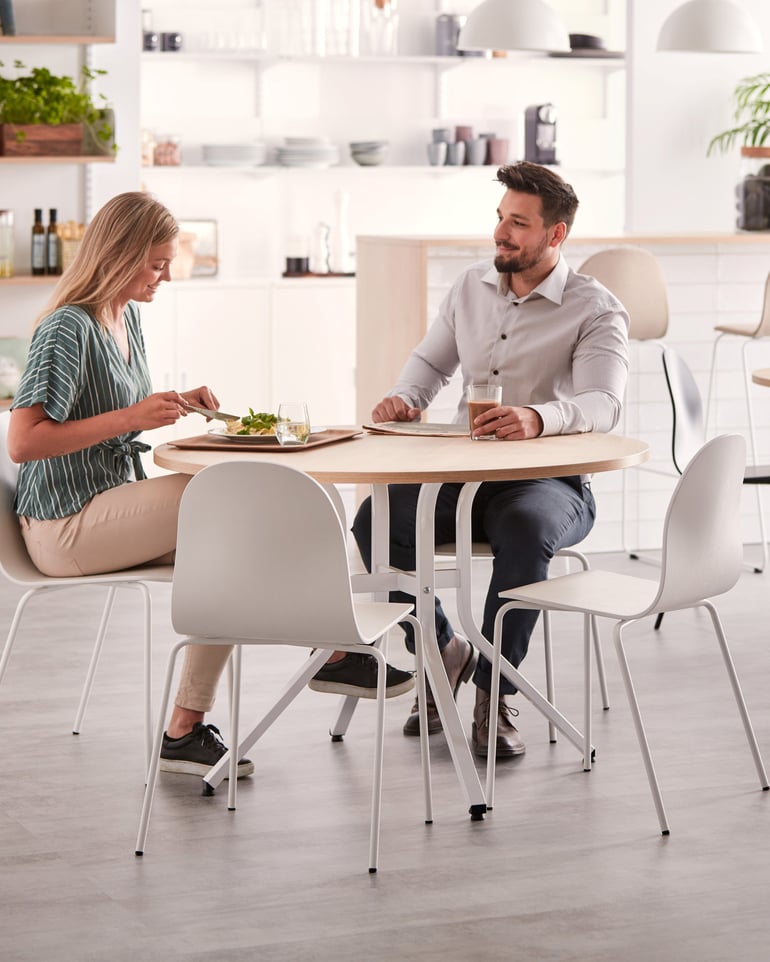 Två personer sitter vid ett runt bord i ett lunchrum med förvaringshyllor i bakgrunden