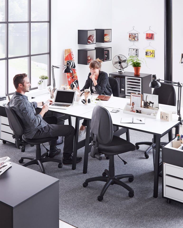 Du darbuotojai kalbasi prie biuro stalo, sėdėdami ant ergonomiškų biuro kėdžių