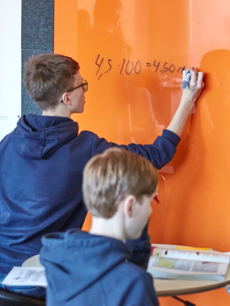 Oppilas kirjoittaa oranssille lasikirjoitustaululle ja luokkatoveri seisoo vieressä