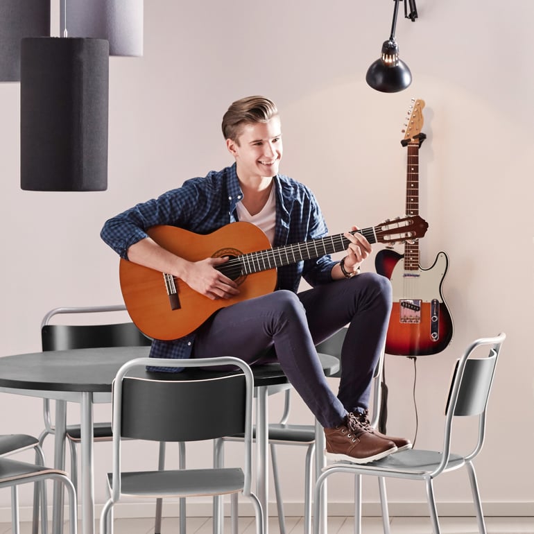 Mokinys su gitara rankose sėdi ant mokyklinio stalo, kojas susidėjęs ant mokyklinių kėdžių