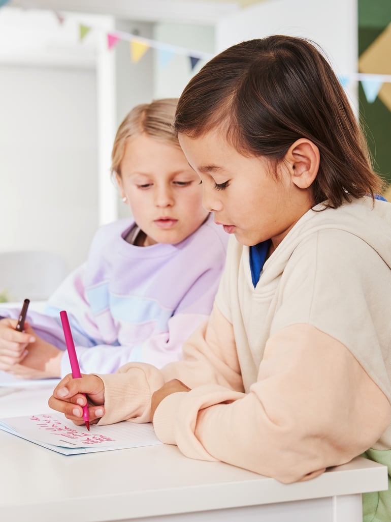 To piger sidder ved et skolebord og skriver på linjeret papir