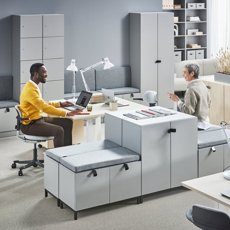 Two people are sitting on office chairs at their desks and talking to each other